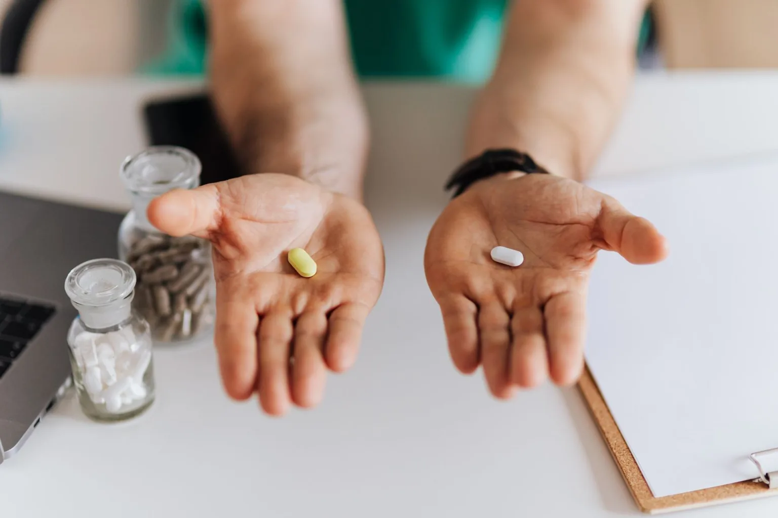 Manos con dos pastillas sobre una mesa de consulta, con un portátil y un portapapeles